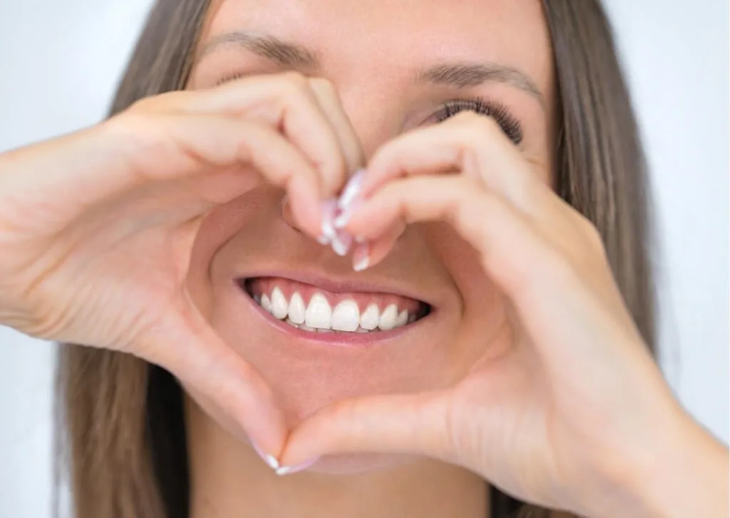 Woman smiling with bright white teeth while making a heart shape with her hands after professional teeth whitening